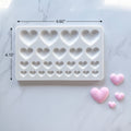 A white rectangular silicone mold with rows of heart-shaped cavities, shown on a light marble surface with three small pink heart-shaped pieces beside it.