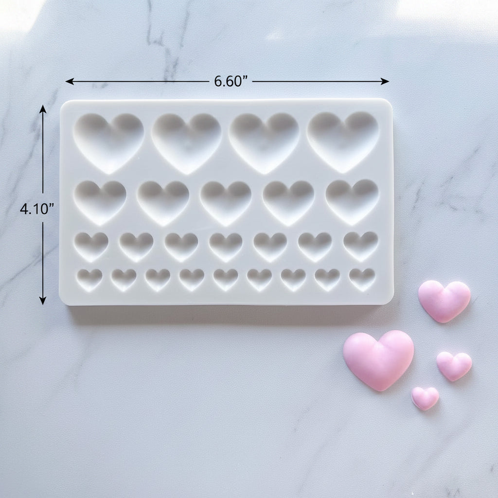 A white rectangular silicone mold with rows of heart-shaped cavities, shown on a light marble surface with three small pink heart-shaped pieces beside it.